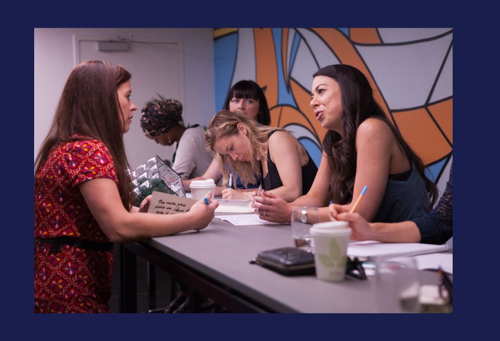 A woman is leading a working session during a workshop. Women are sitting along a table writing in their workbooks.