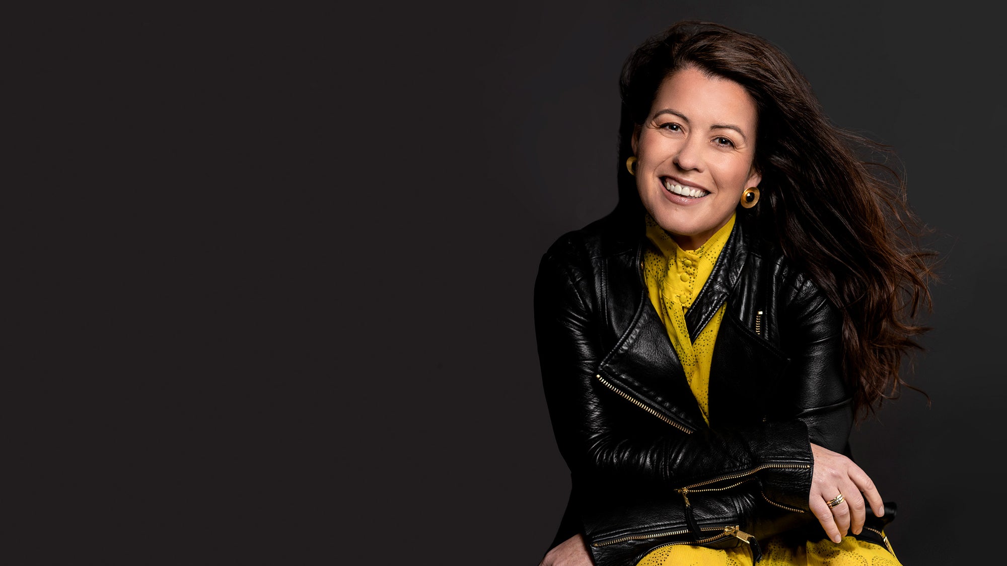 Woman in black leather jacket and yellow dress smiling and sitting, looking at the camera.