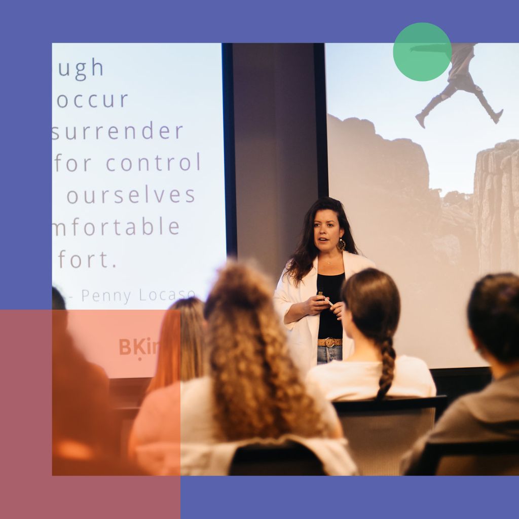 A woman is speaking to an audience in front of projector screens with her slideshow.