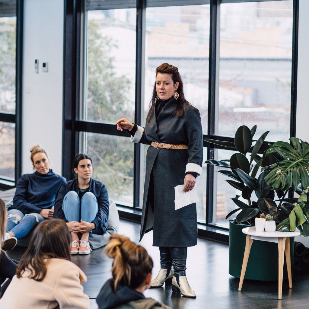 A woman is standing and speaking to an audience that are sitting comfortable on the floor.