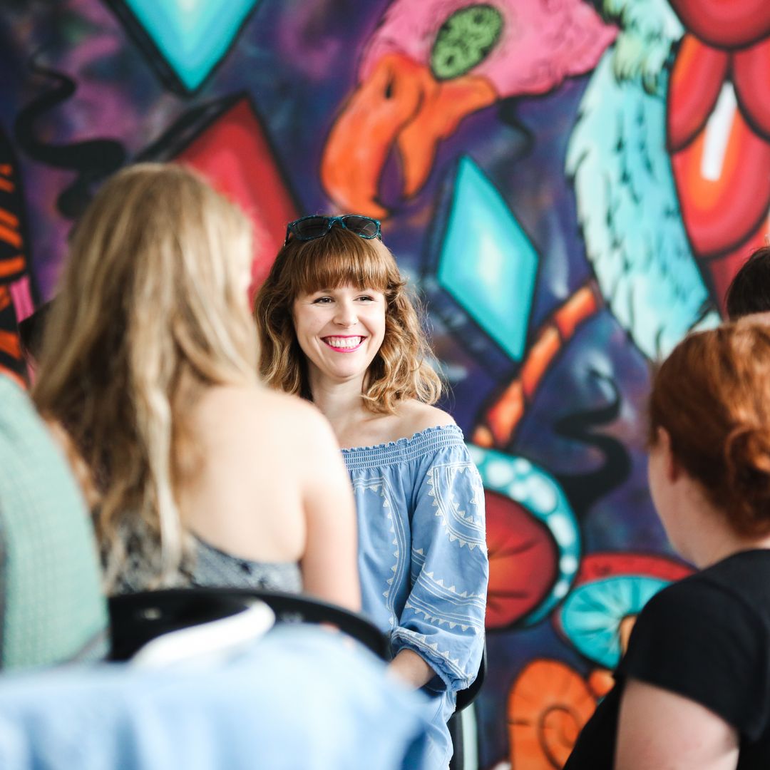 A woman is smiling and engaged in a conversation with two other women in front of colorfully painted wall.